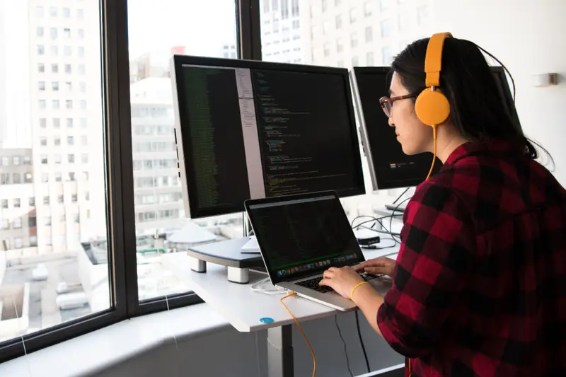 Mujer trabajando con dos monitores y un portátil junto a una ventana